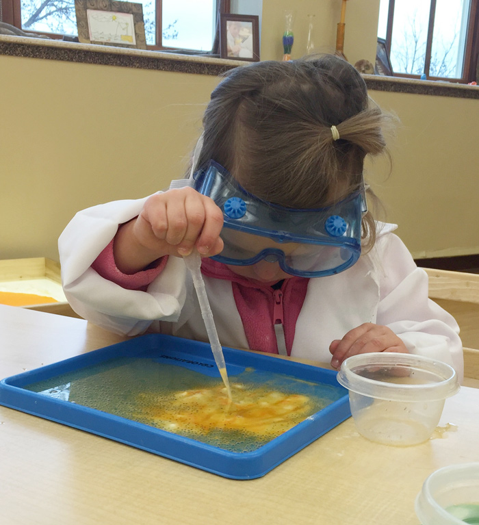 Toddler exploring fizzy bubbles experiment with pipette wearing science goggles.