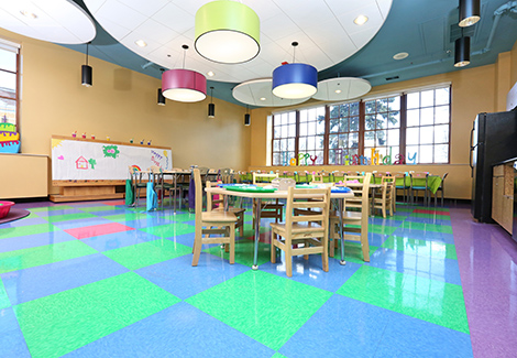 Wooden chairs and tables set up with activities in brightly lit and colorful room with large windows.