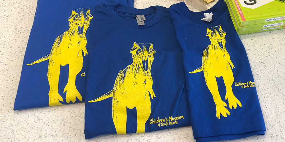 The gift shop has a fresh stock of blue and yellow Mama T. Rex shirts. 