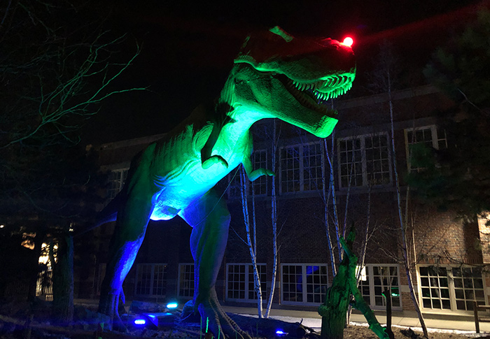 T. rex bathed in green light with a glowing red nose standing outside the Children's Museum of South Dakota. 