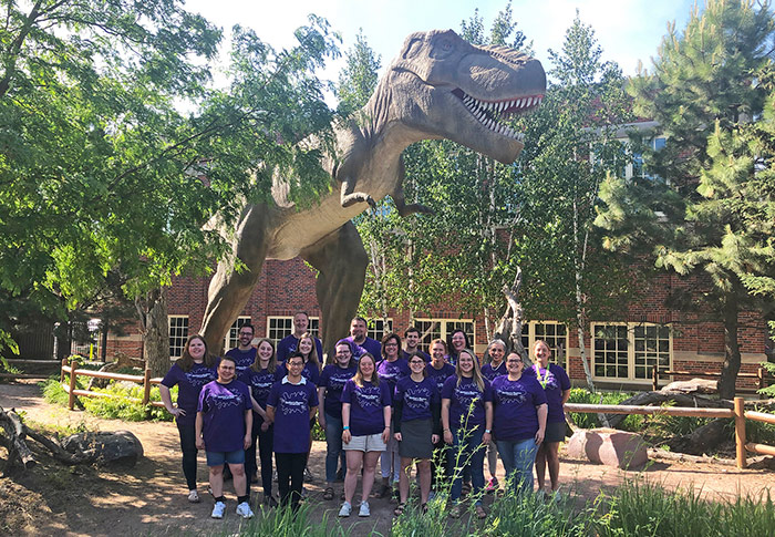 Group of staff members of Children's Museum of South Dakota in purple tshirts standing in front of animatronic lifesize T. rex.
