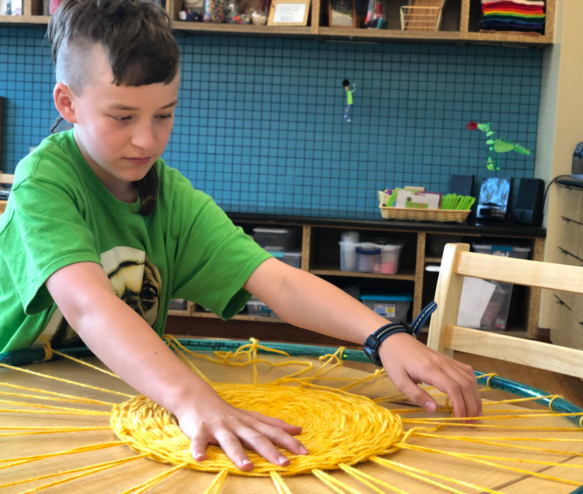 Boy weaving yellow yarn using a hula hoop as a frame sitting at a wooden table in an art studio