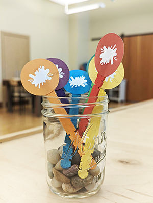 Rainbow colored popsicle sticks with circles taped to them placed in a clear glass canning jar with pebbles in the bottom. 