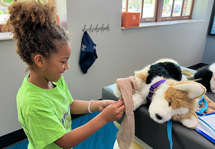 Girl bandaging a stuffed animal puppy who is laying on a vet office table