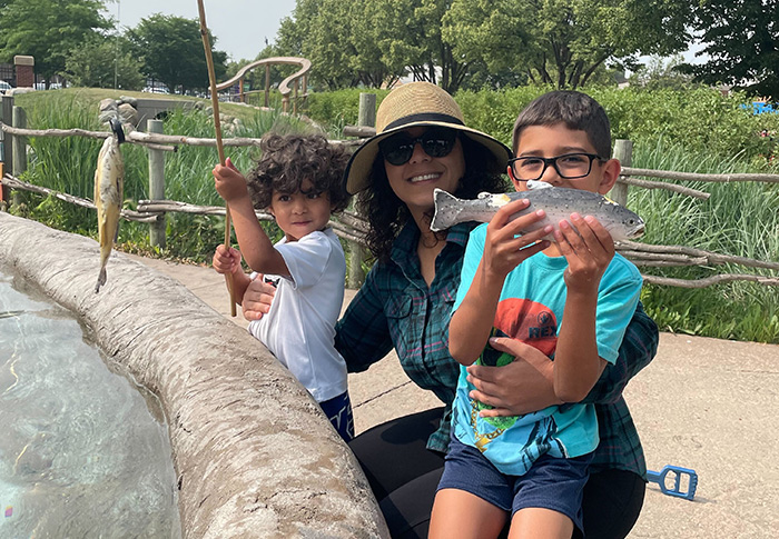 Woman in sun hat and sunglasses kneeling with two young boys, one standing with bamboo fishing pole and second holding a lifelike replica of a fish.