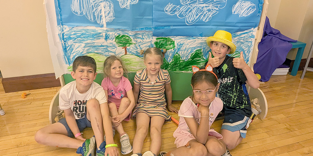 Five children sitting in front of a hand-drawn backdrop of clouds and trees smiling at the camera.