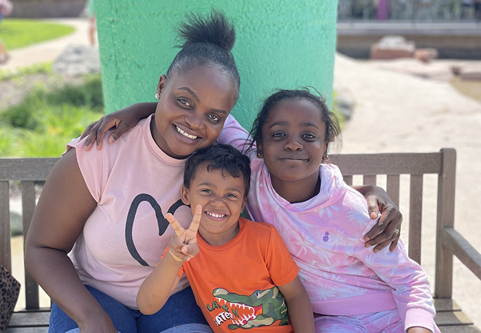 Family of 3 sitting on a bench outside and smiling, middle family member gives the peace sign.