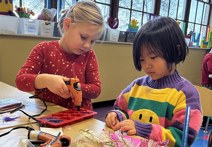 Two girls working on craft projects with hot glue gun. They are in a natural looking room with bright windows.