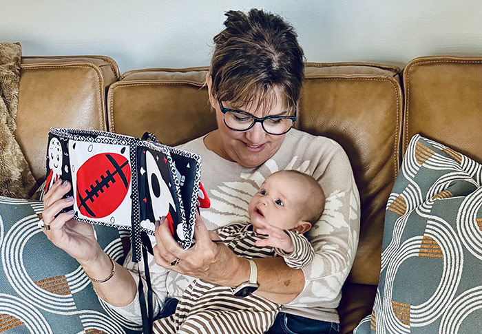 Woman with glasses is sitting on a leather sofa holding up a book and holding an infant in a striped onesie.