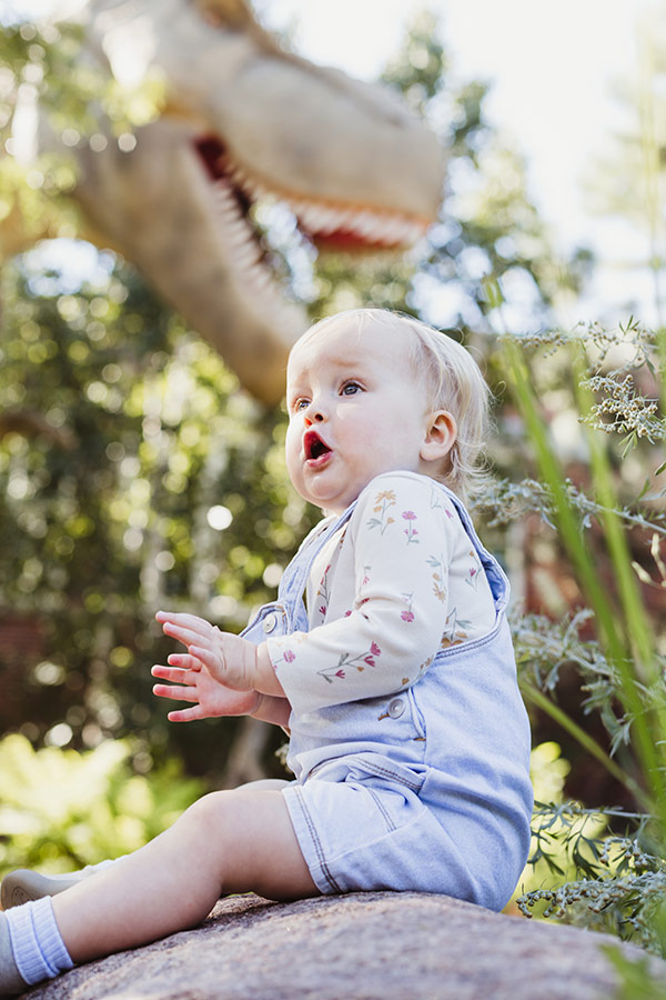 Little girl sitting on a rock in front of a animatronic T. rex with a ROAR expression on her face. 