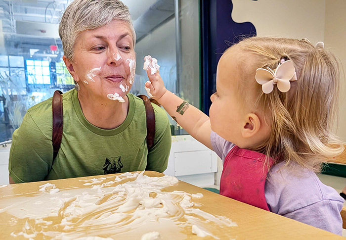 Little girl and grandma sit in front of a table covered in shaving cream. The little girl is adding shaving cream to the grandma's face. 