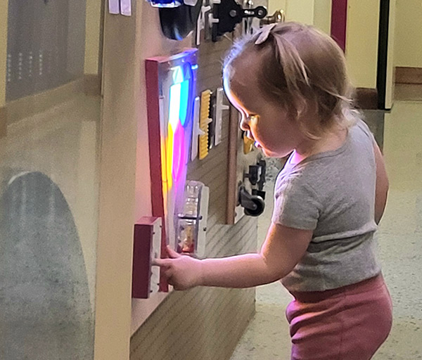 Little girl standing and exploring on the tinker wall --- a wall of switches, lights, locks, and knobs.